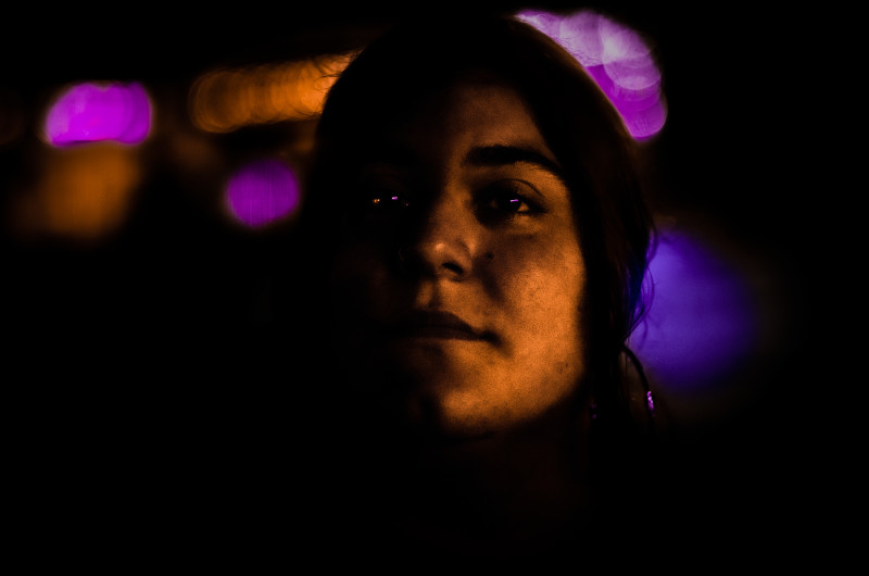 a girl partly in shadow looks upward at the camera, purple lights flaring behind her and reflected in her eyes, while an orange light glows on the right side of her face