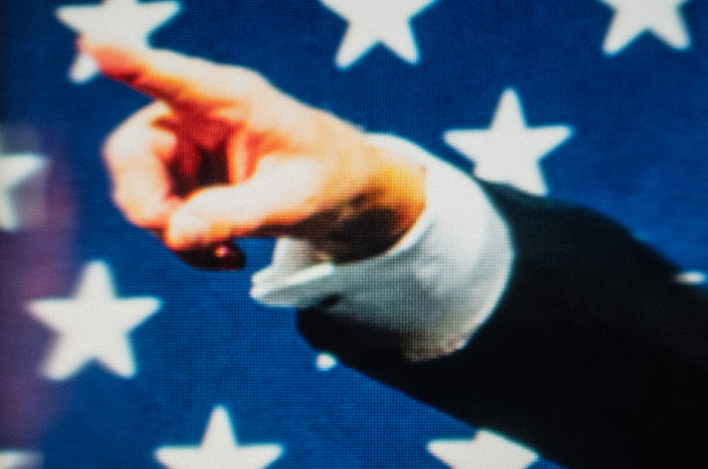 in front of the stars of the US Flag, a man's hand points straight up to the left