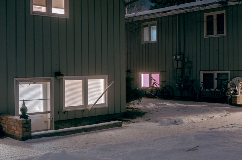 two green paneled apartment buildings in winter, at night, in snow. a basement apartment emits a pink glow