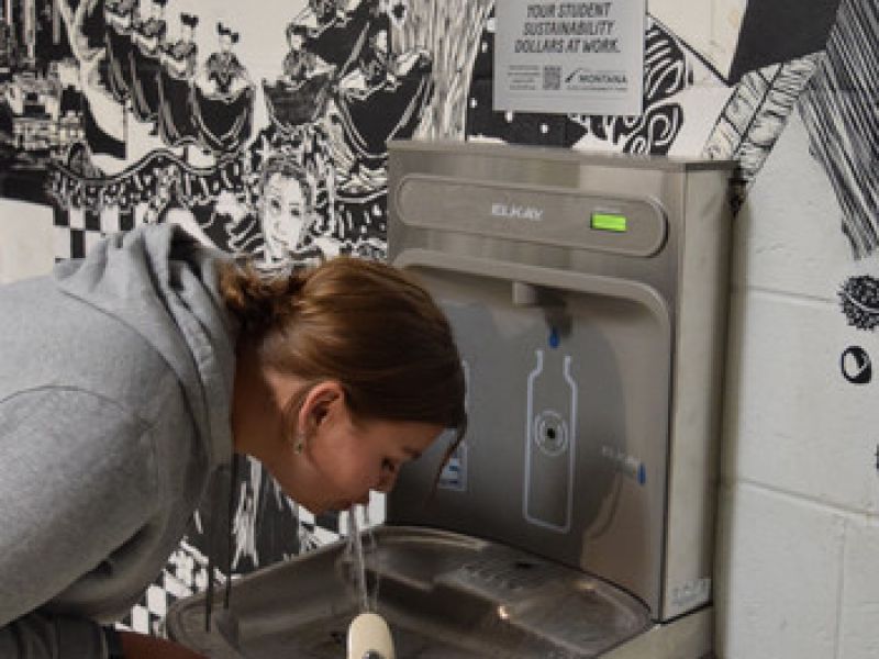 Zoe drinks from the new drinking fountain with Kless sign above it. Wall has a mural background of random drawings