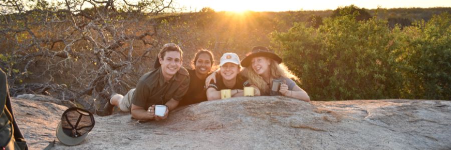 four people with coffee cups laying on top of a large rock with sun coming up behind them
