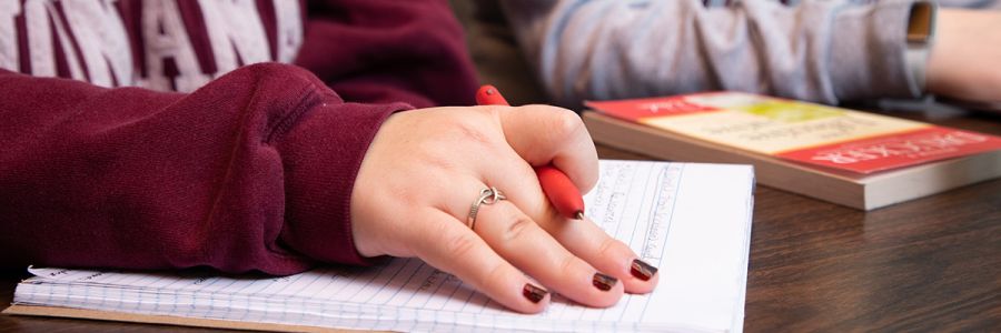 close up view of a hand holding a pen over a notebook with books in the background