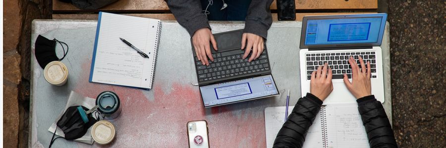 view from above of two students working on laptops at a table covered in books, cups of coffee and more