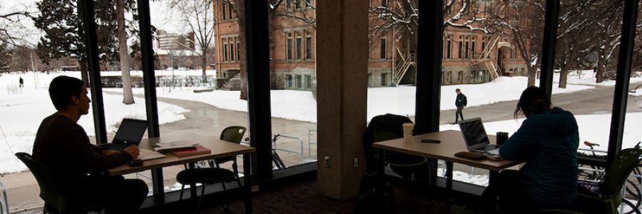 silhouettes of two students studying at tables in front of windows with a view of the snowy UM campus