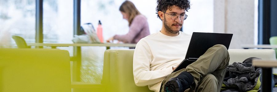 student sitting and working on a laptop with students studying at tables in the background