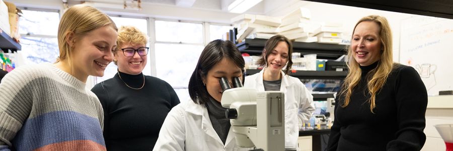 student looking into a microscope with four other students watching behind them
