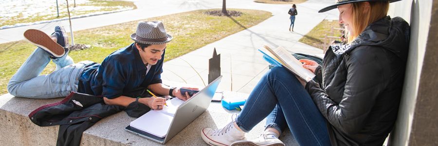 one student reading a book sitting against a building while another lays in front of them working on a laptop