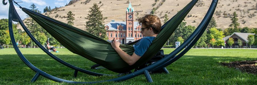 student reading outside in a hammock on a sunny day with university hall in the background