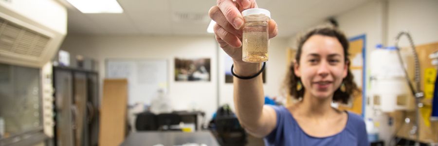 student in a lab holding a jar up to the camera containing a liquid filled with debris