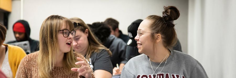 two students in a classroom talking to each other with various students in the background