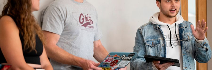 three students holding tablets at the front of a classroom while one of them speaks