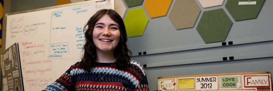 smiling person standing in front of their laptop with a whiteboard and other displays behind them