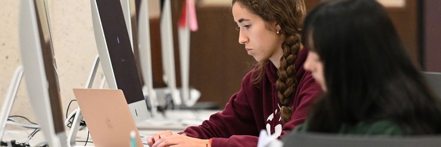 student working on a laptop alongside a row of desktop computers on a table
