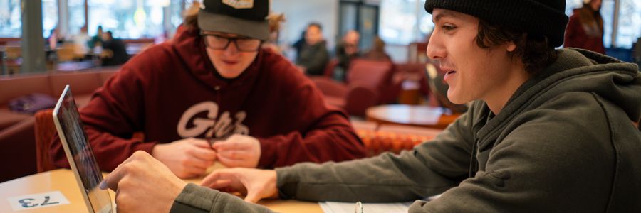 two students sitting at a table looking at a laptop while one points at something on the screen