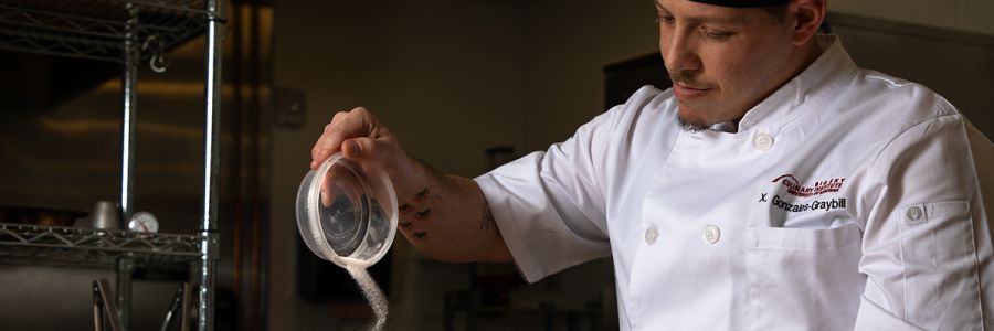 chef in a kitchen pouring spices from a bowl into another container not shown in the image