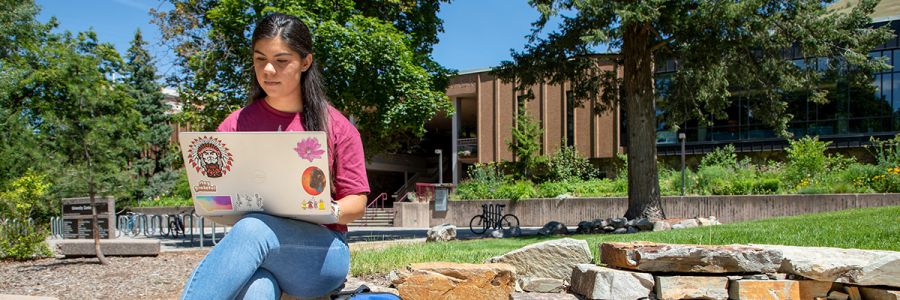student working on laptop outside on a sunny day with the university center in the background
