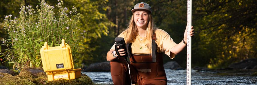 student wearing wadders in a creek with scientific instruments and a large stick for depth measurement