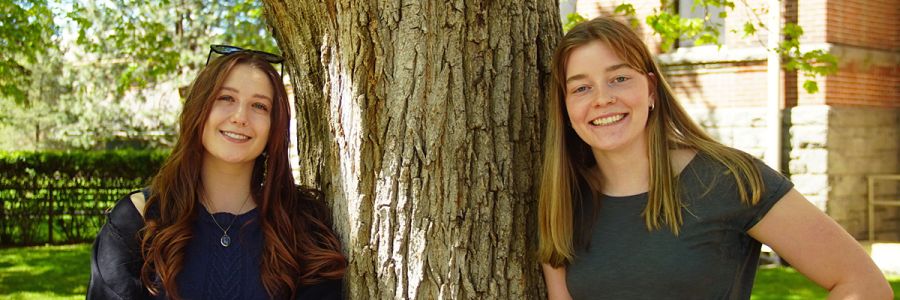 two people smiling and posing outside while leaning against a tree