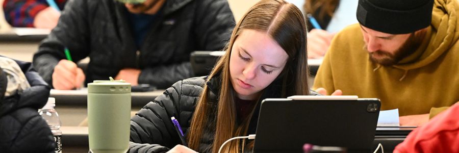 student taking notes next to a laptop with rows of other students behind them