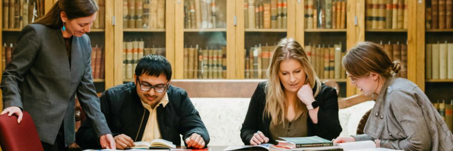group of four people reading books and studying at a table with cabinets full of books in the background