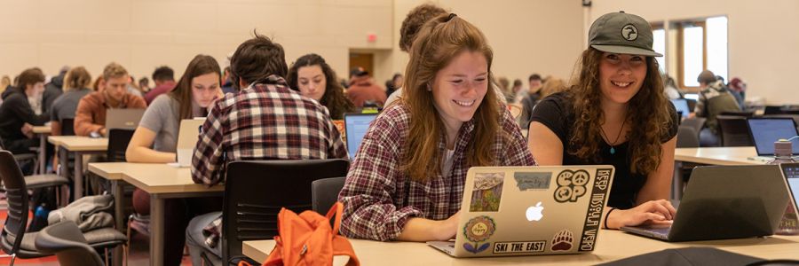 two students working on laptops sitting at a table with many more tables of students working in the background