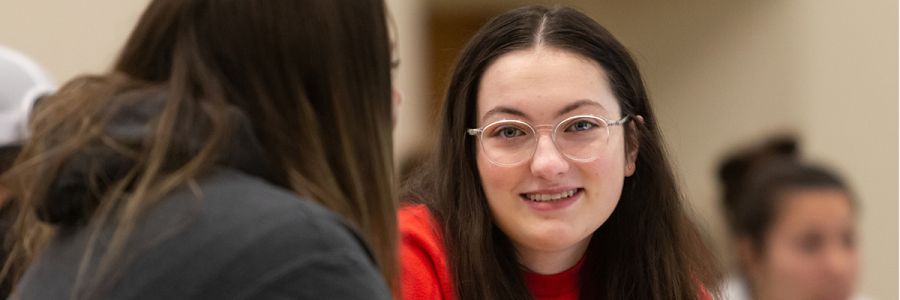 two students talking while sitting at a table with one student looking into the camera