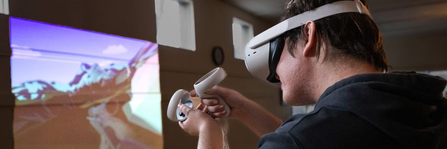 student wearing a virtual reality headset and holding controllers pointed at an image projected on a classroom wall