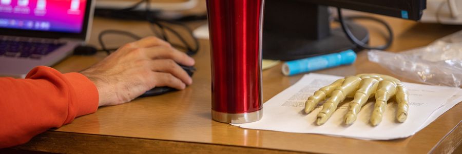 desk with a hand holding a mouse next to a model of the bones of a human hand resting on some papers
