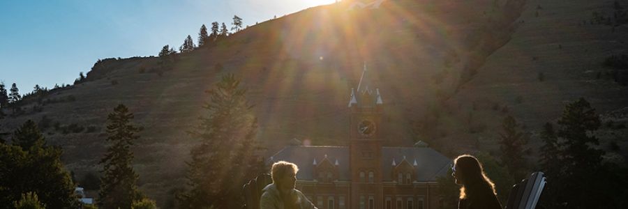 two students sitting on the UM oval talking with university hall in the background and the sun dramatically shining from behind mount sentinel