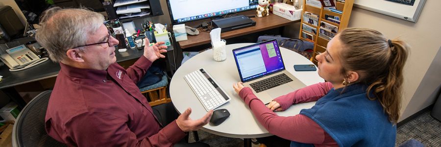 two people working at a table one talking and another in front of a laptop