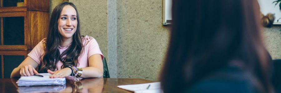 student sitting at table smiling with hands holding a large note filled notebook