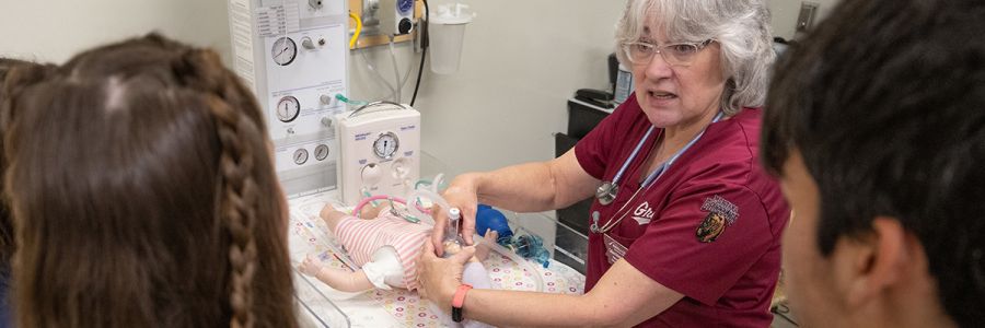 two students watching a nurse work on a training dummy of a baby