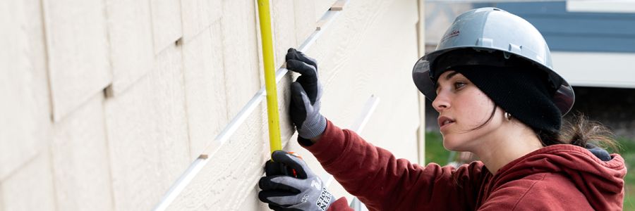 student in construction safety gear measuring the side of a building with measuring tape