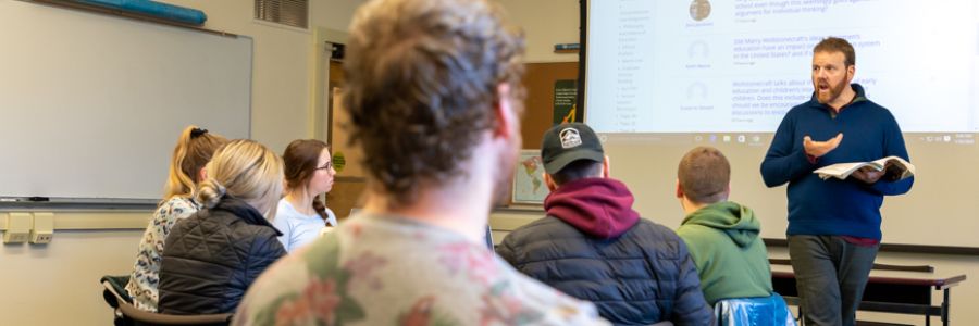 professor walking and talking with open book in their hands in front of a classroom while students watch and listen