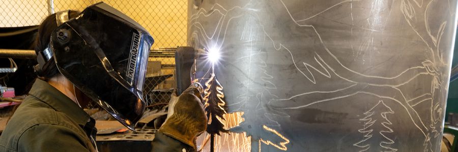 student wearing welding mask while cutting a tree image out of a large piece of metal