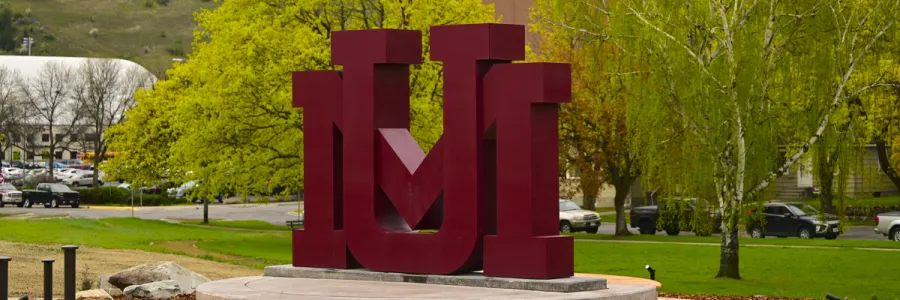 Maroon intertwined block letter "U" and "M" sculpture on a concrete base near the Madison street bridge close to the 91Ԫ campus in Missoula, MT with grass and trees in the background.