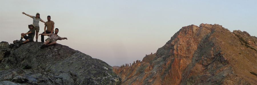four people posing on top of a rocky mountain peak with another peak in the background