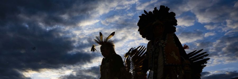 silhouettes of two people in tradition Native American clothing in front of a dramatic sky