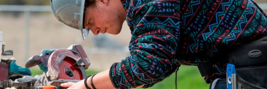 A picturesque view of University Hal featuring a clock tower and green spire, surrounded by lush trees with autumn foliage. The campus is set against the backdrop of rolling hills covered in dry grass and patches of forest, under a bright blue sky with scattered clouds.