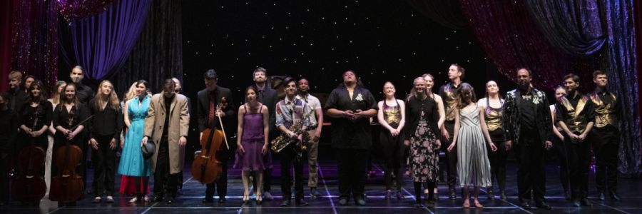 large group of performers standing on a darkened stage with a starry background