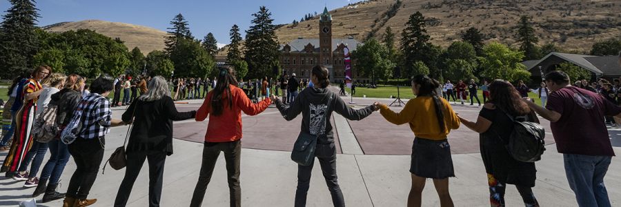 large group of students holding hands around the center of the UM oval