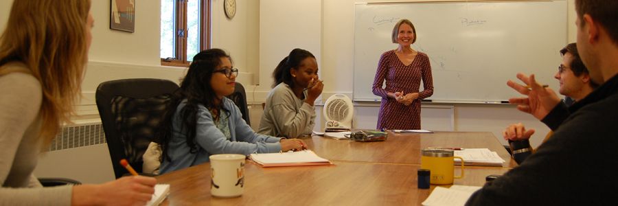 table of students talking with professor standing at the front by a whiteboard