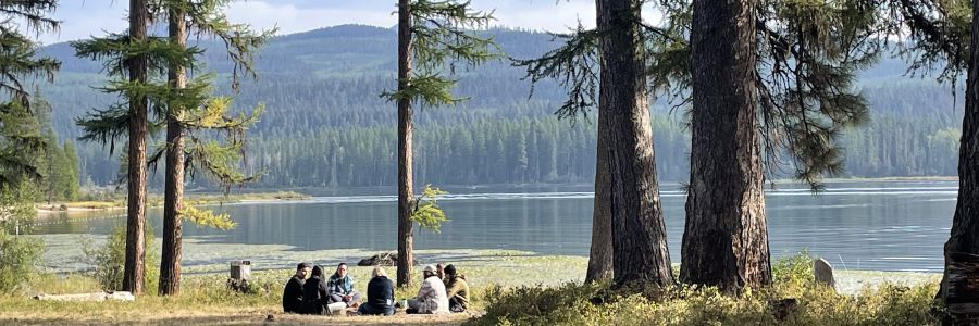people sitting in a circle on the shore of a lake with conifer tree covered hills in the background