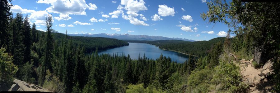 a lake seen from a tree covered mountain with more mountains in the distance