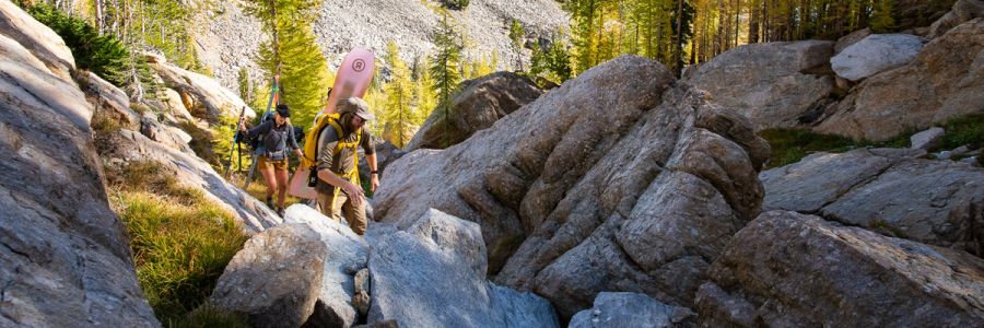 two people hiking through large rocks with a snowboard and skis on their backs