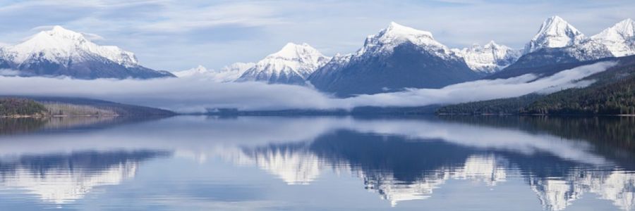 steep snow covered mountains with fog below them being reflected in a still lake