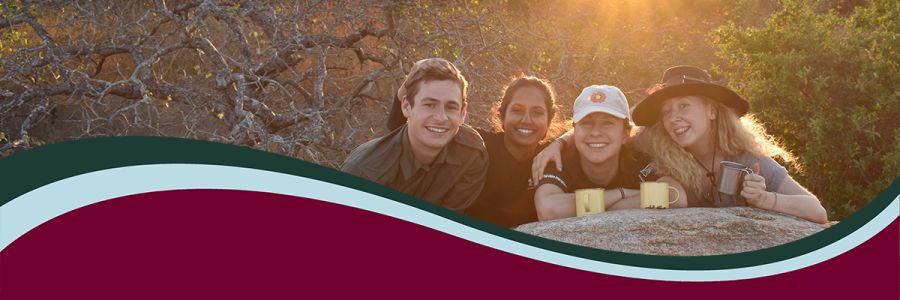 four students posing with coffee cups and leaning on a rock with sun shining behind them