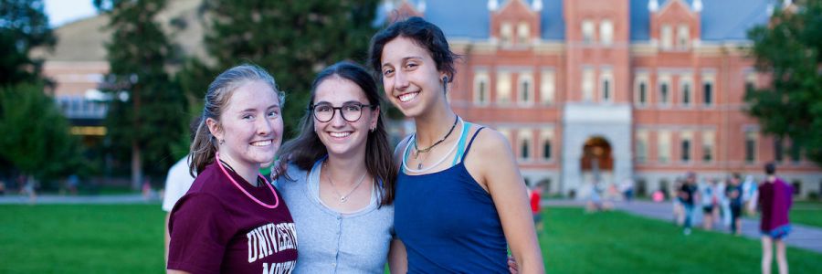 three students smiling and posing together on the oval with University Hall in the background