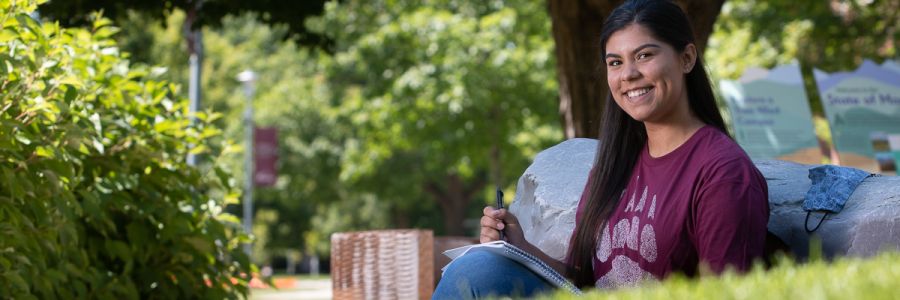 smiling student outside sitting against a rock with a notebook and pen