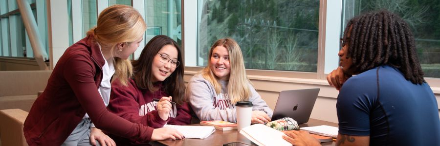 four students with books and a laptop at a table listening to one of them talk